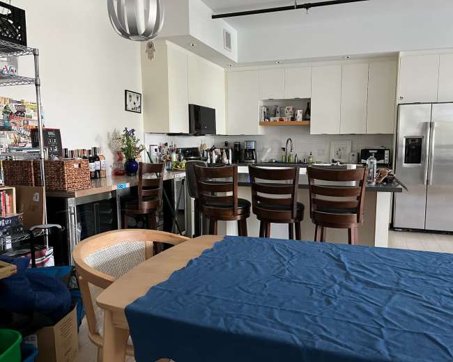 A modern kitchen and dining area features a wooden dining table with a blue tablecloth, bar stools at a kitchen island, and shelves with various kitchen supplies and bottles.