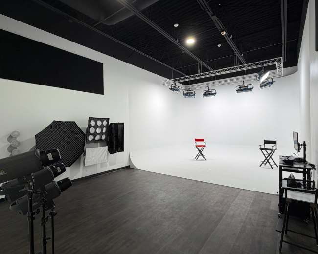 The image shows a photography studio with two director's chairs, various lighting equipment, and a plain white backdrop.
