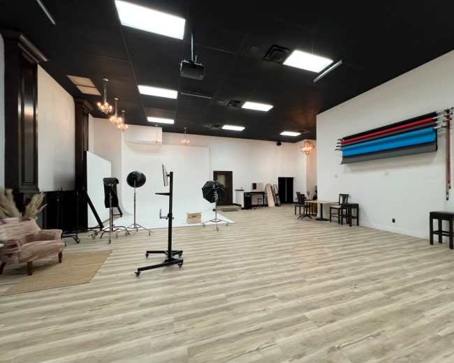 The image shows a spacious photography studio with wooden flooring, various lighting equipment, and colorful backdrops hanging on the wall.