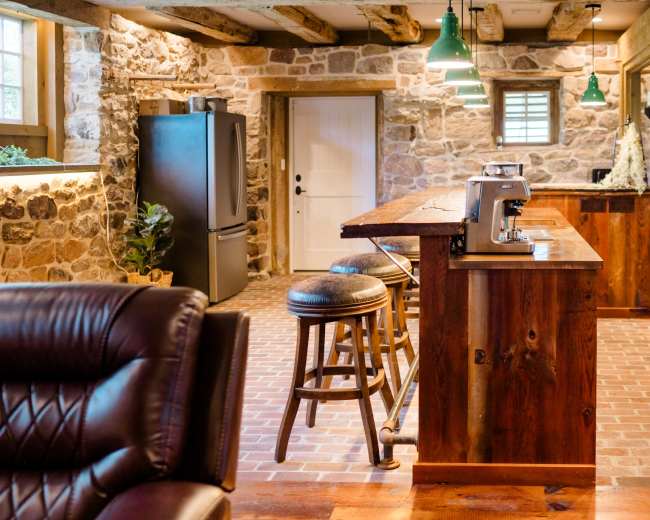 The image depicts a rustic kitchen with stone walls, wooden beams, and a bar area featuring stools and modern appliances.
