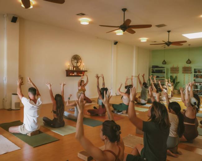 A group of individuals is practicing yoga in a spacious studio, with some seated on mats and others engaged in various poses while raising their arms.