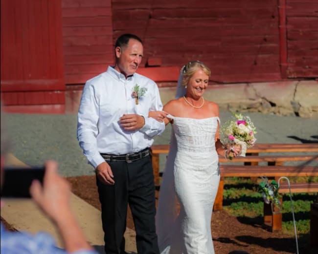 A bride in a white gown walks down a path alongside a man, while holding a bouquet and her dog walks beside them.