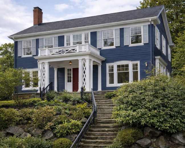 The image shows a two-story blue house with white trim, a red front door, and landscaped greenery surrounding stone steps leading to the entrance.