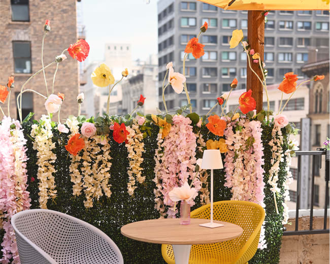 The image shows a small outdoor seating area with a white table and two chairs—one gray and one yellow—set against a backdrop of colorful artificial flowers.