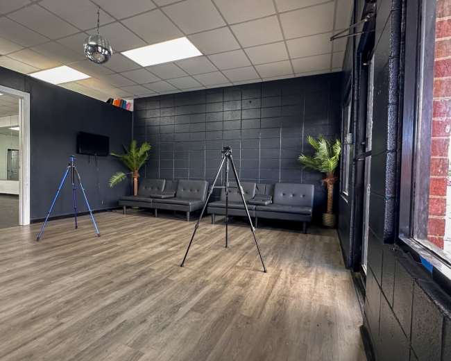 The image shows a spacious room with dark walls, two tripods set up in front of a row of gray sofas, and a large window allowing natural light.