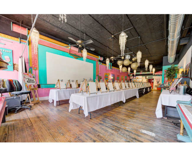 The image shows an art studio setup with multiple easels and blank canvases arranged along a row of tables in a colorful, well-lit space.
