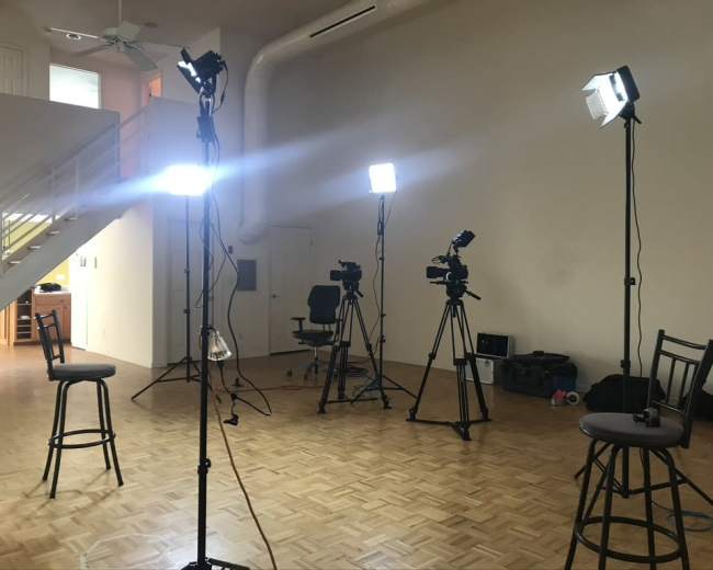 A well-lit recording studio with multiple camera setups and bar stools arranged in a spacious, empty room.