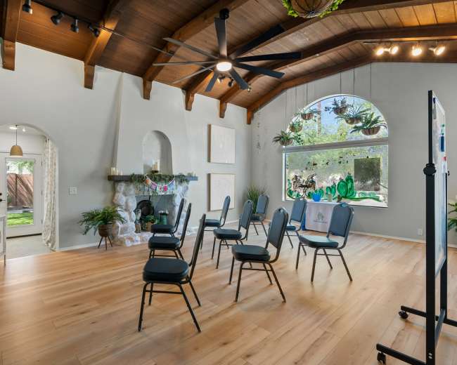 The room features wooden beams on the ceiling, a stone fireplace, and a circular window filled with plants, with several chairs arranged in a semicircle.