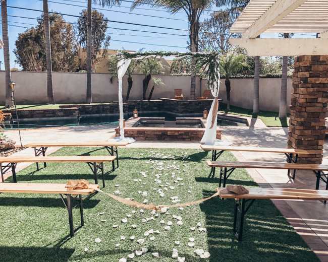 A wedding setup features a stone altar surrounded by benches and a path of scattered flower petals leading to it, all set in a backyard with a swimming pool and palm trees.