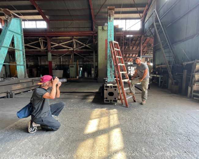 A person is crouching and taking a photograph of another person who is standing on a ladder in a spacious industrial setting.
