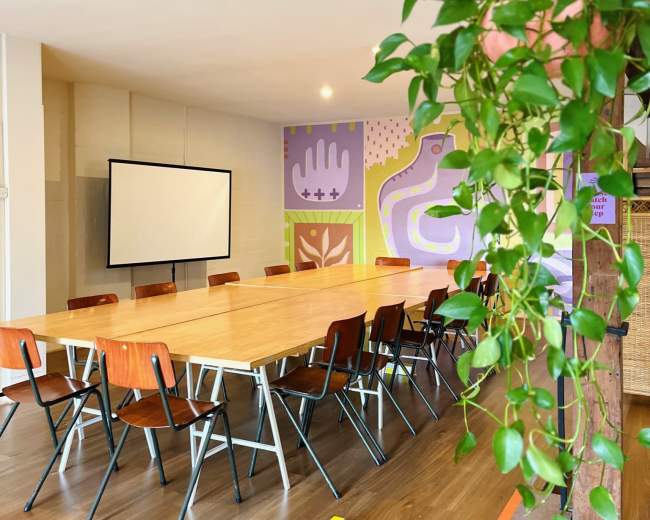 The image shows a spacious meeting room with a long wooden table surrounded by chairs, featuring colorful wall murals and a projector screen at the front.