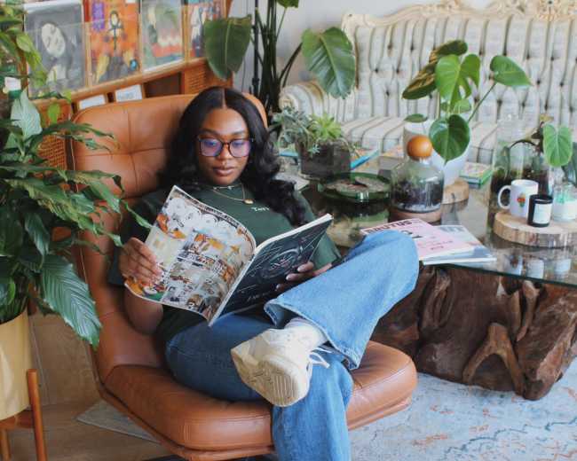 A person sits comfortably in a modern chair, reading a magazine in a cozy room filled with plants and decorative furniture.
