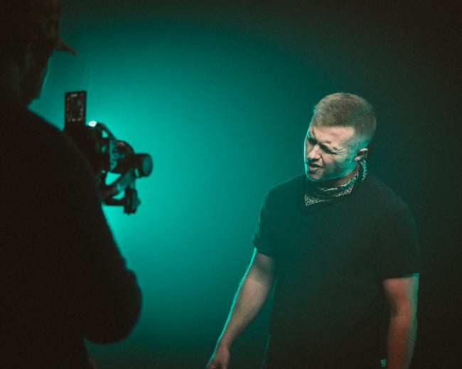 A man in a black shirt and bandana performs in front of a camera with blue-green lighting in a studio setting.