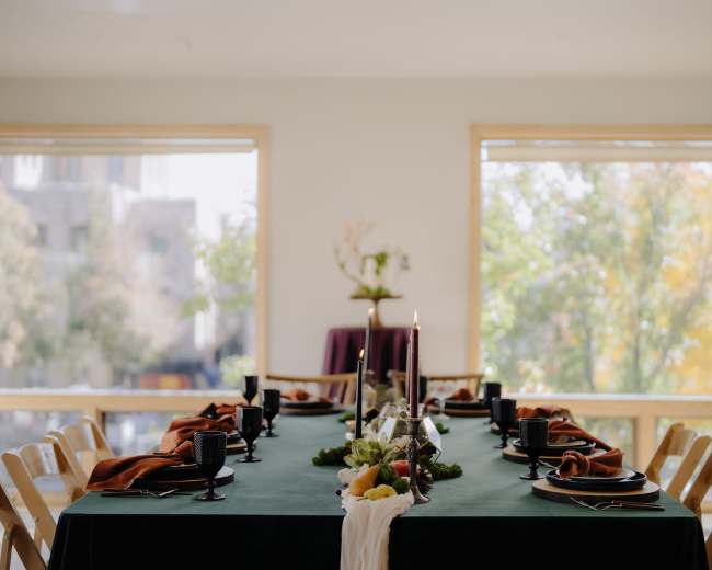 A long dining table is set with dark green tablecloth, black glassware, and decorative elements for a meal, surrounded by chairs, with large windows letting in natural light and views of autumn foliage outside.