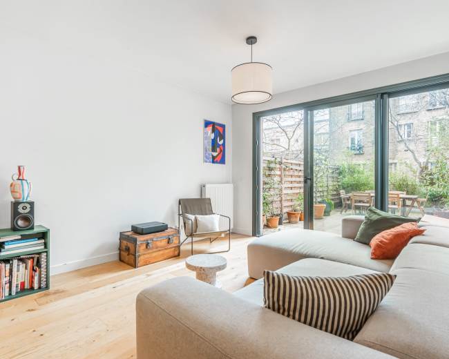 A modern living room features a light-colored sofa, a wooden floor, and large glass doors opening to a garden area.