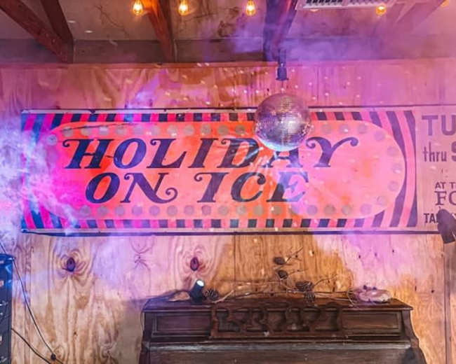 A large sign reading "Holiday on Ice" is displayed on a wooden wall, illuminated by colorful lights and surrounded by smoke effects.