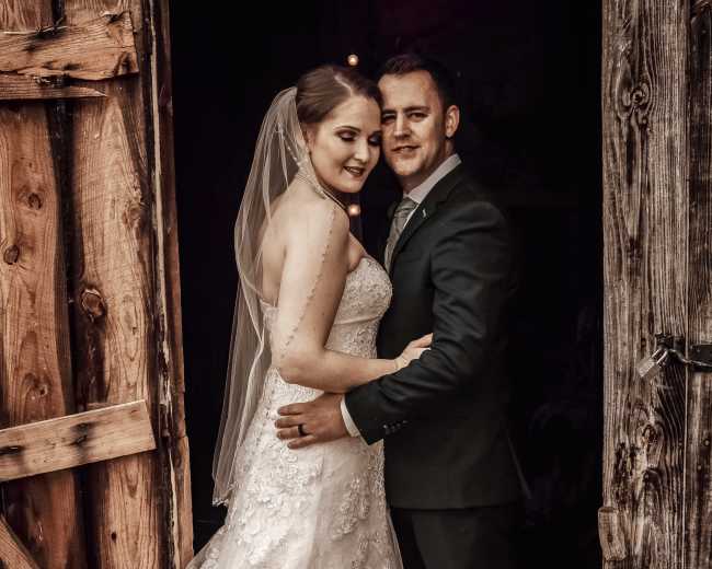 A bride in a white lace gown and a groom in a suit pose together at an entrance of a rustic wooden building.