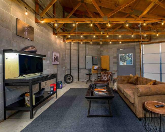 The image shows a spacious, industrial-style room with a couch, a coffee table, a television, and overhead string lights.