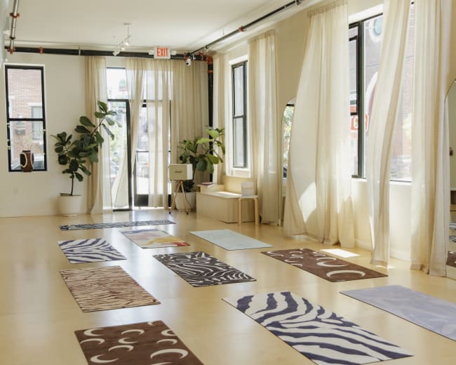 The image shows a bright yoga studio with several colorful mats arranged on the floor and large windows allowing natural light to enter.