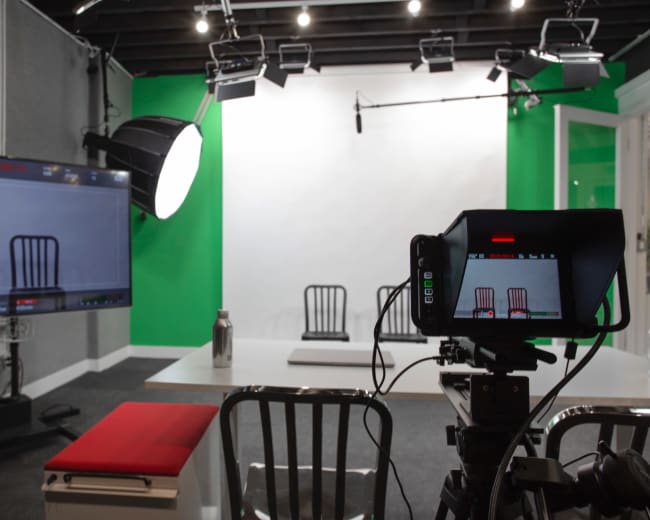 A video recording studio equipped with a camera, a large monitor, and several chairs arranged in front of a plain backdrop.