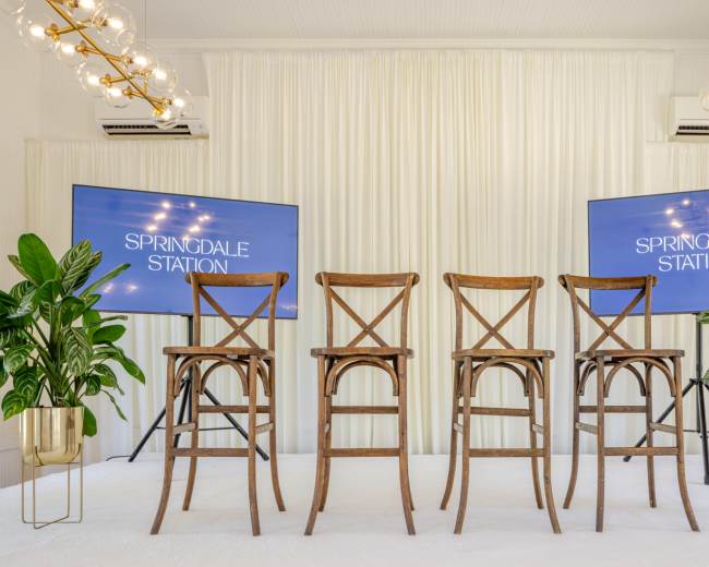 The image shows a staged area with four wooden bar stools in front of two large screens displaying the text "SPRINGDALE STATION," surrounded by potted plants.
