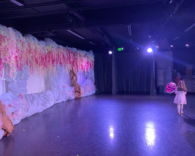 A stage is set with fabric drapes and floral arrangements, while a girl in a dress stands in the spotlight.