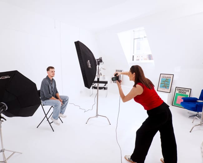 A photographer is capturing a portrait of a man sitting on a chair in a bright studio equipped with professional lighting equipment.
