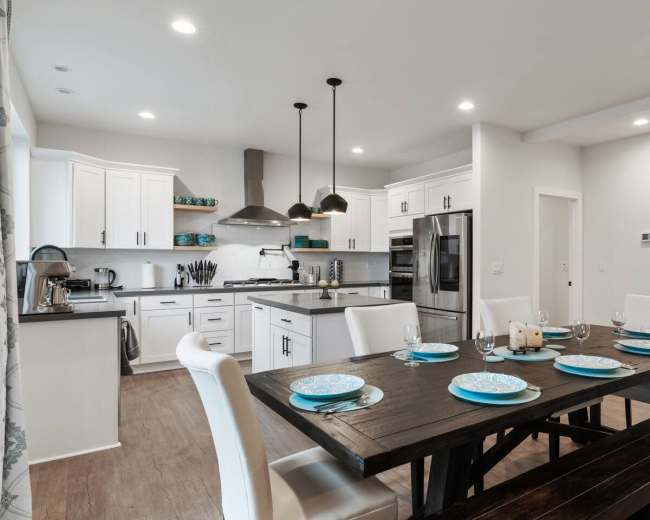 A modern kitchen and dining area features a large dark wood table set with blue dishware, surrounded by white chairs, and equipped with white cabinetry and stainless steel appliances.
