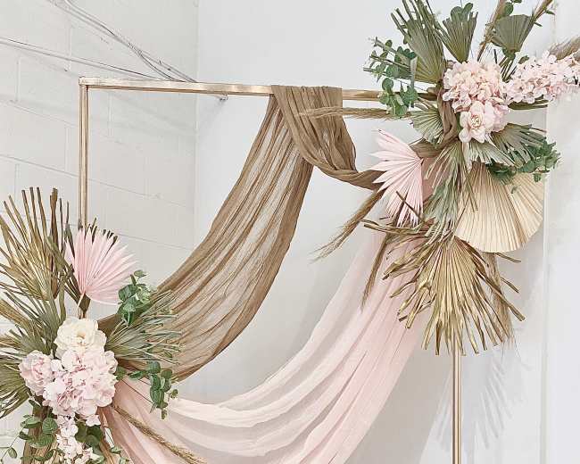The image shows a decorative backdrop featuring draped fabrics in soft pink and neutral tones, accented with floral arrangements and palm leaves.