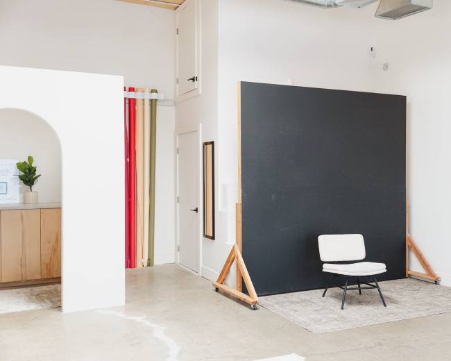 The image shows a minimalist interior space featuring a black wall-mounted panel, a white chair on a rug, and a wall with red and beige vertical stripes in the background.