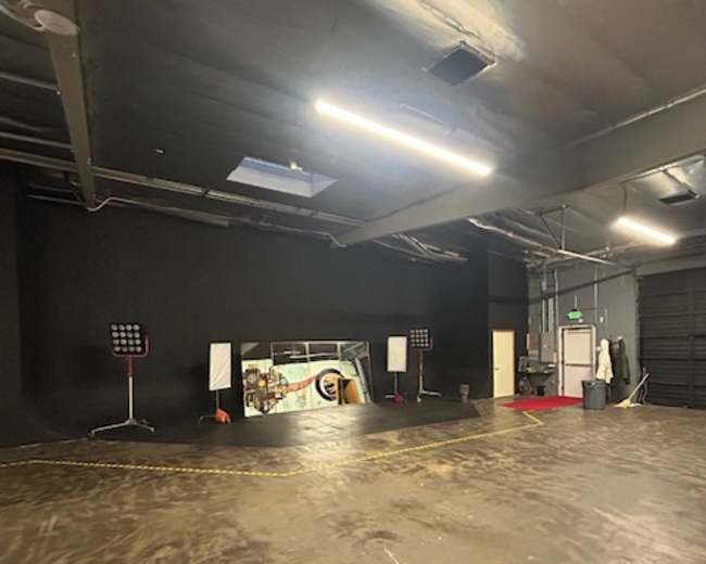 The image shows a spacious black-walled industrial studio with a polished concrete floor, featuring stage lights and a large mirror at the far end.