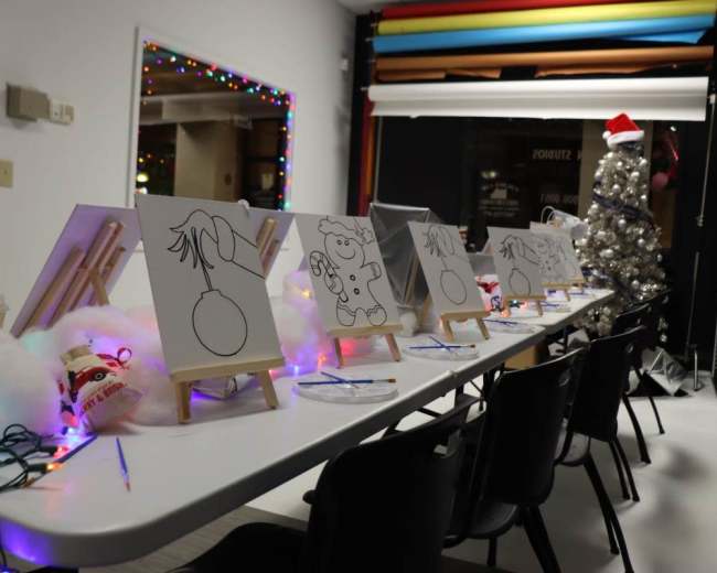 A long table is set for a painting activity, with blank canvases on easels and colorful decorations, including lights and a holiday-themed tree.