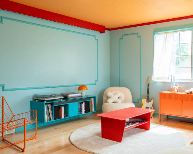 The room features a light blue wall with decorative trim, a bright orange ceiling, and colorful furniture including a red coffee table, orange cabinet, and a white chair.