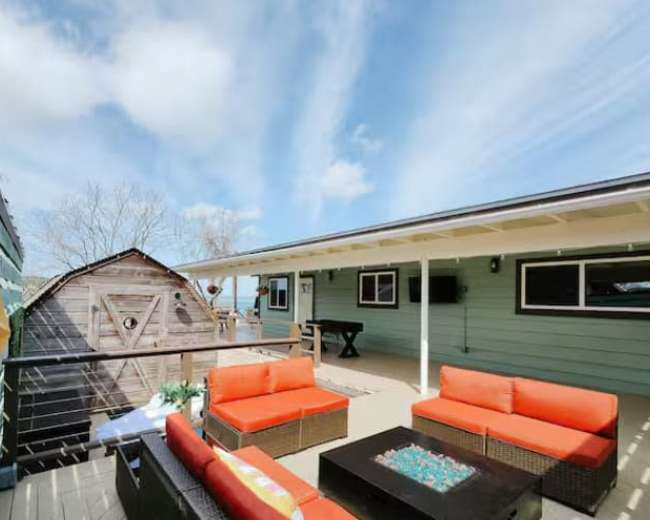 The image shows a spacious outdoor deck with orange seating and a fire pit, surrounded by wooden structures and a clear blue sky.