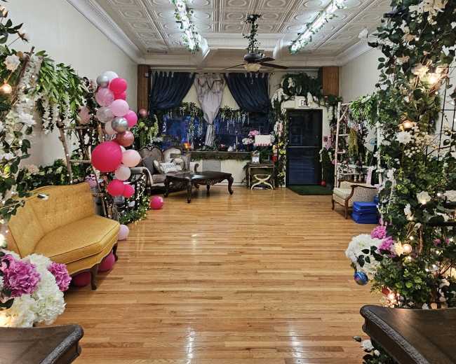 The image shows a decorated indoor space with wooden flooring, featuring floral arrangements and balloons along the walls, and seating areas with vintage furniture.