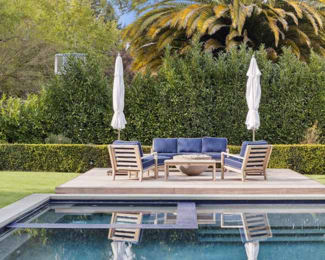 A wooden deck features a seating area with blue cushions and a fire pit, surrounded by a neatly trimmed hedge and palm trees, with a swimming pool reflecting the scene.