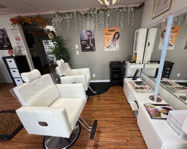 The image shows a hair salon interior with two white styling chairs, a mirror, and various magazines on a table, all under decorative greenery and posters on the walls.