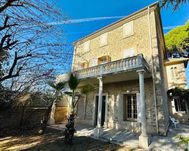 A three-story stone house with a balcony and columns stands in a garden with trees and clear blue skies.
