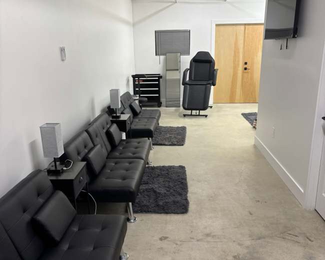 The image shows a minimalist waiting area with five black leather couches along one wall, grey rugs on the concrete floor, and a reception desk at the end of the hallway.