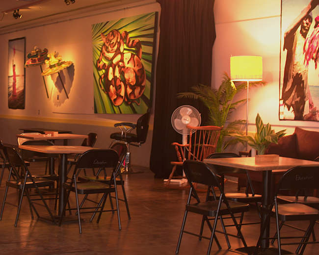 The image shows an empty interior space featuring several tables and chairs, with colorful artwork displayed on the walls and a small stage area in the background.