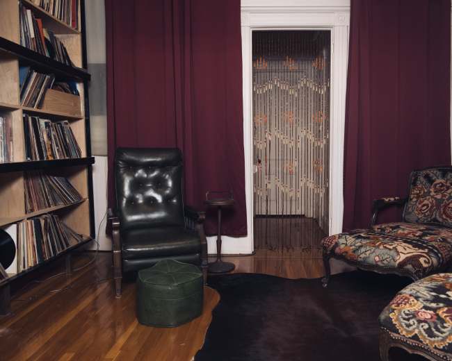 A cozy living room features two chairs, a small table, and a bookshelf filled with vinyl records, all framed by maroon curtains and a patterned screen door.