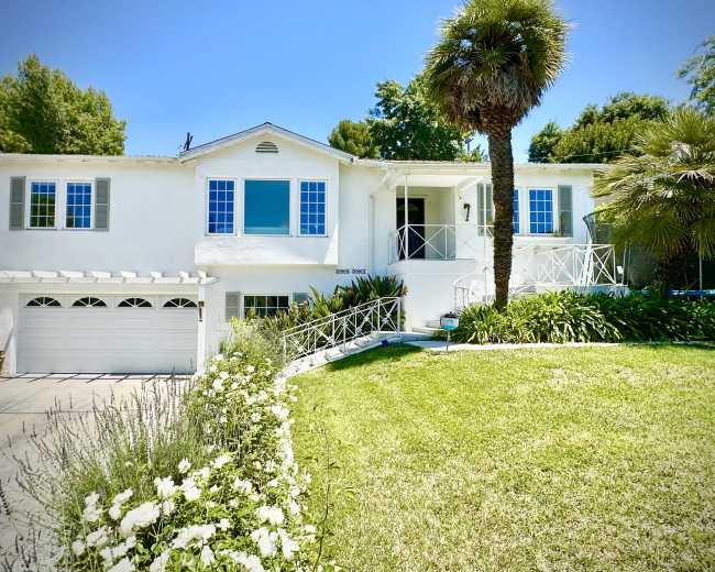 A white house with a front staircase is surrounded by greenery and flowers, featuring a two-car garage on the left.