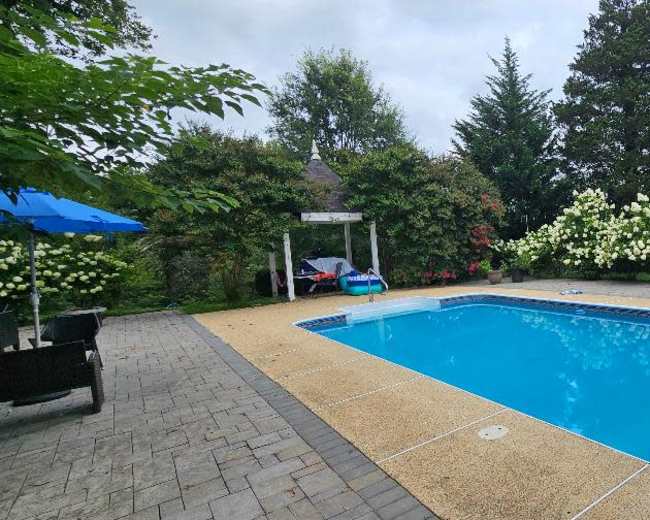 A rectangular swimming pool surrounded by flower-filled hedges and a stone patio, with garden furniture and an umbrella nearby.