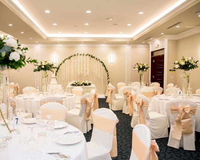 The image shows a beautifully arranged banquet hall with round tables covered in white linens and adorned with light-colored bows, featuring floral centerpieces and a decorative backdrop.