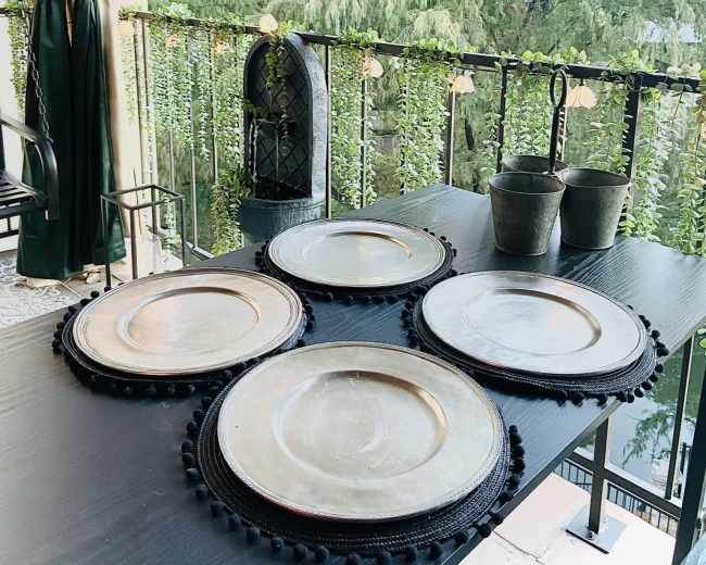 A set of round, metallic plates is arranged on a dark table with greenery and buildings visible in the background.