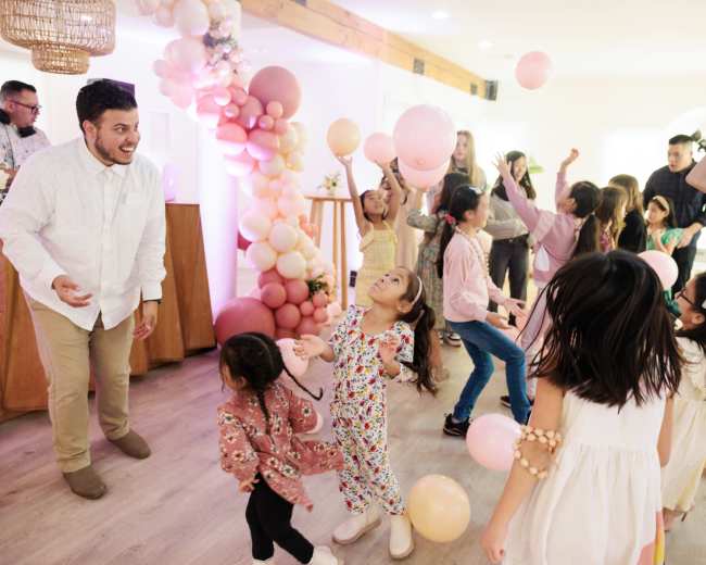 A group of children and adults dance joyfully at a celebration, surrounded by colorful balloons and decorations.