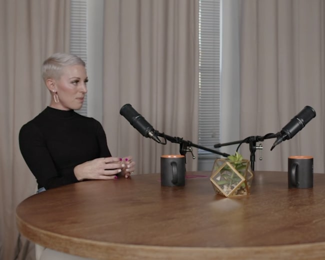 Two women are seated at a round wooden table with microphones and small decorative items, engaged in conversation.