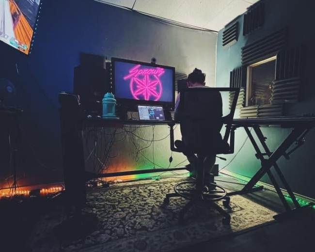 A person sits at a desk with a computer and colorful lighting in a dimly lit room that features soundproofing panels.