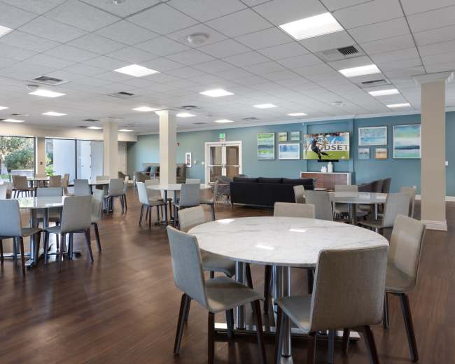 The image shows a spacious dining area with multiple round tables, chairs, and large windows letting in natural light.