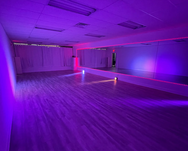 A dance studio is illuminated with purple and red lighting, featuring wooden flooring and a large mirror on one wall.